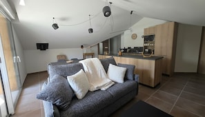 Ein modernes Wohnzimmer mit einem grauen Sofa, einer Küchenzeile mit Holzschränken und einem Esstisch.