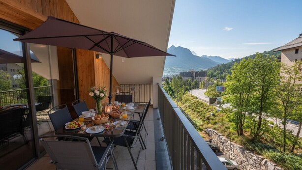 Un balcone con un tavolo apparecchiato per la colazione, sedute all'aperto e vista su una catena montuosa.
