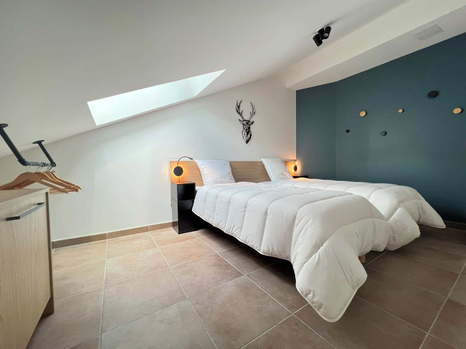 A bedroom with a skylight, a bed with a white comforter, a wall-mounted deer head decoration, and a blue accent wall.