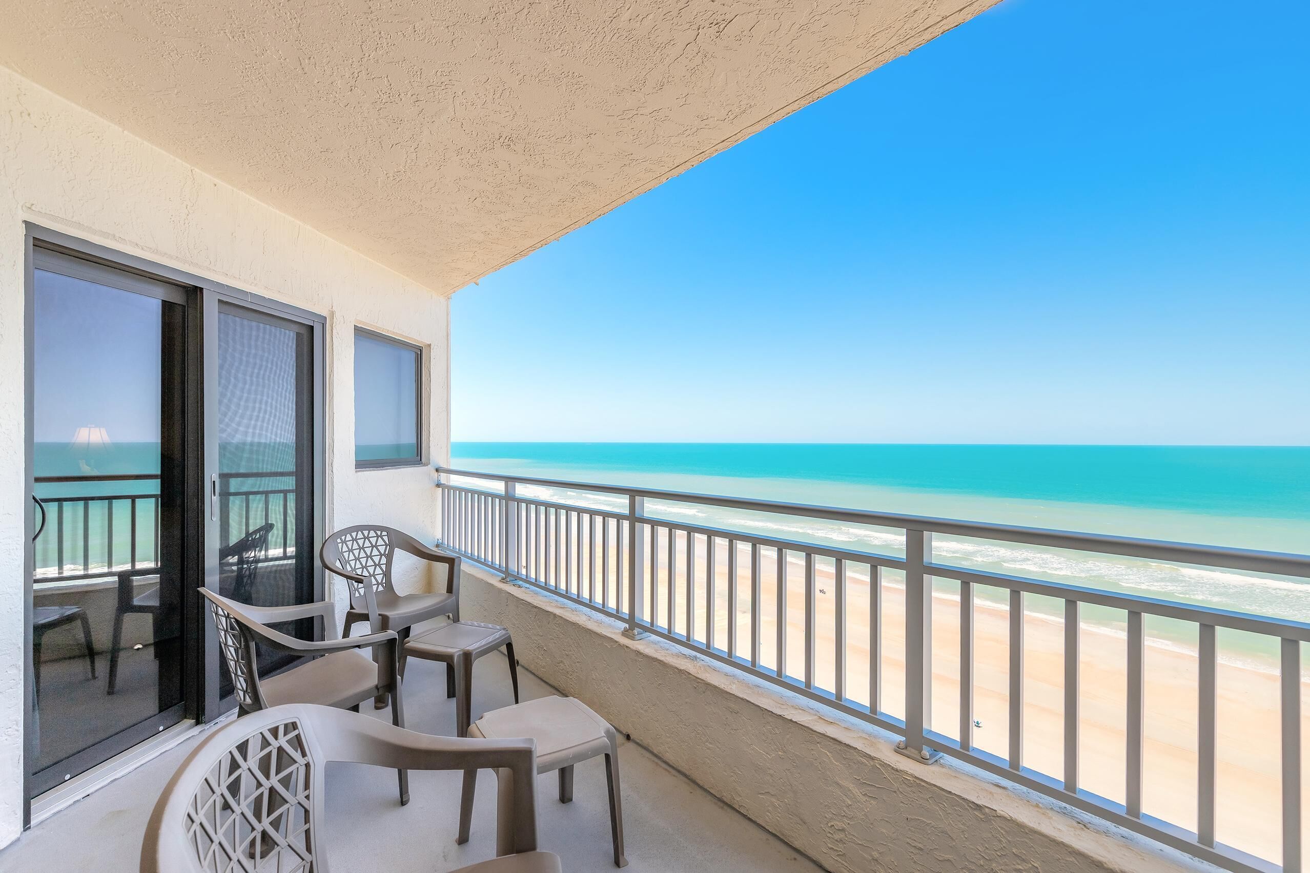 A balcony with a view of the beach, a table, chairs, and a glass door.