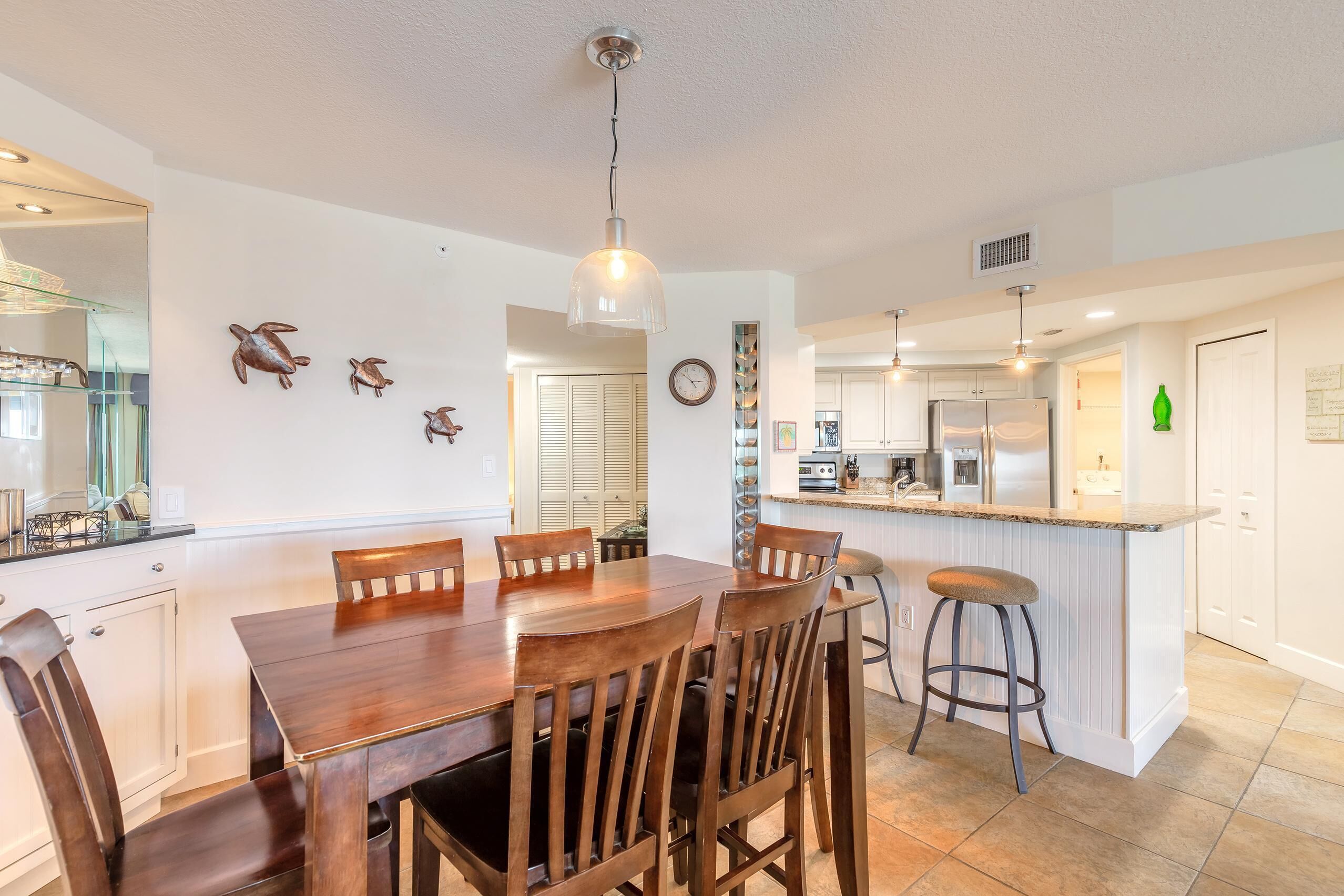 A kitchen with a central island, wooden dining table, and chairs. There are wall-mounted decorative items and a clock.