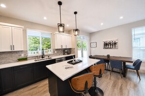 A modern kitchen with a central island, bar stools, a dining table, and pendant lighting. - The Blue Pearl - Oyhut Bay Seaside Resort (Ocean Shores)