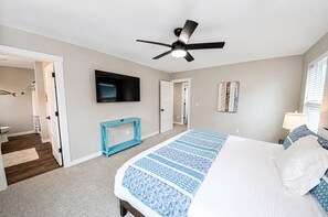 A bedroom with a bed, a ceiling fan, a TV, a console table, and a hallway leading to a bathroom. - The Blue Pearl - Oyhut Bay Seaside Resort (Ocean Shores)