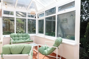 A conservatory with multiple windows, green cushioned chairs, and a white table. - Gorway Cottage (Teignmouth)