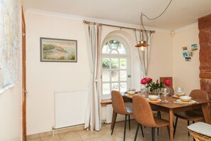 A dining area with a table set for four, a window with curtains, a radiator, and a painting on the wall. - Gorway Cottage (Teignmouth)