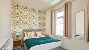 Un dormitorio con cama, mesita de noche, radiador, ventana con cortinas y papel tapiz floral en la pared.