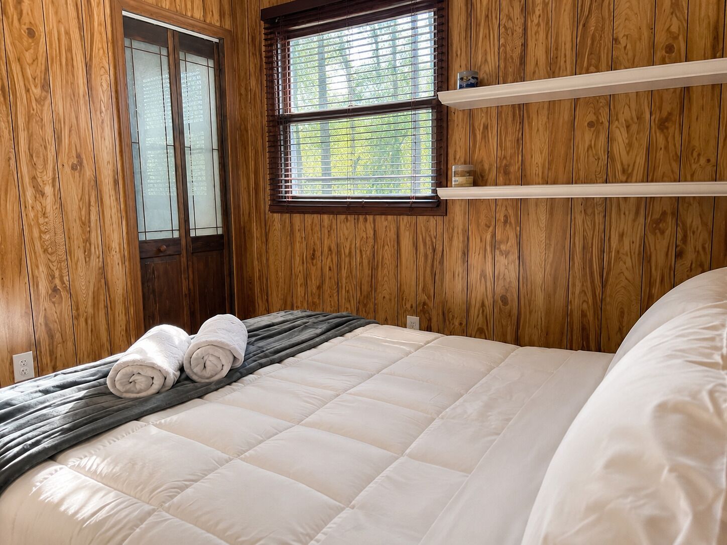 Een kamer met houten lambrisering, een bed met een geborduurde matras, twee opgerolde handdoeken, een raam met jaloezieën en een plank boven het bed.