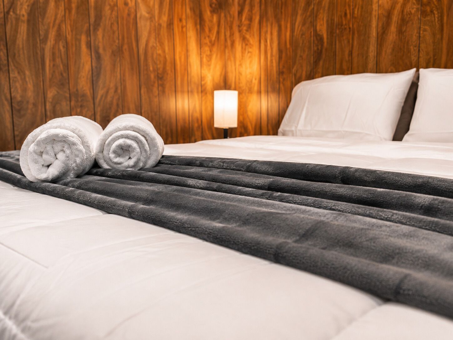 Una cama bien hecha con sábanas blancas, una manta gris y dos toallas blancas enrolladas.