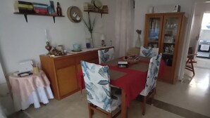 Uma sala de jantar com uma mesa posta para quatro pessoas, um armário de madeira com portas de vidro e uma pequena mesa com toalha rosa.