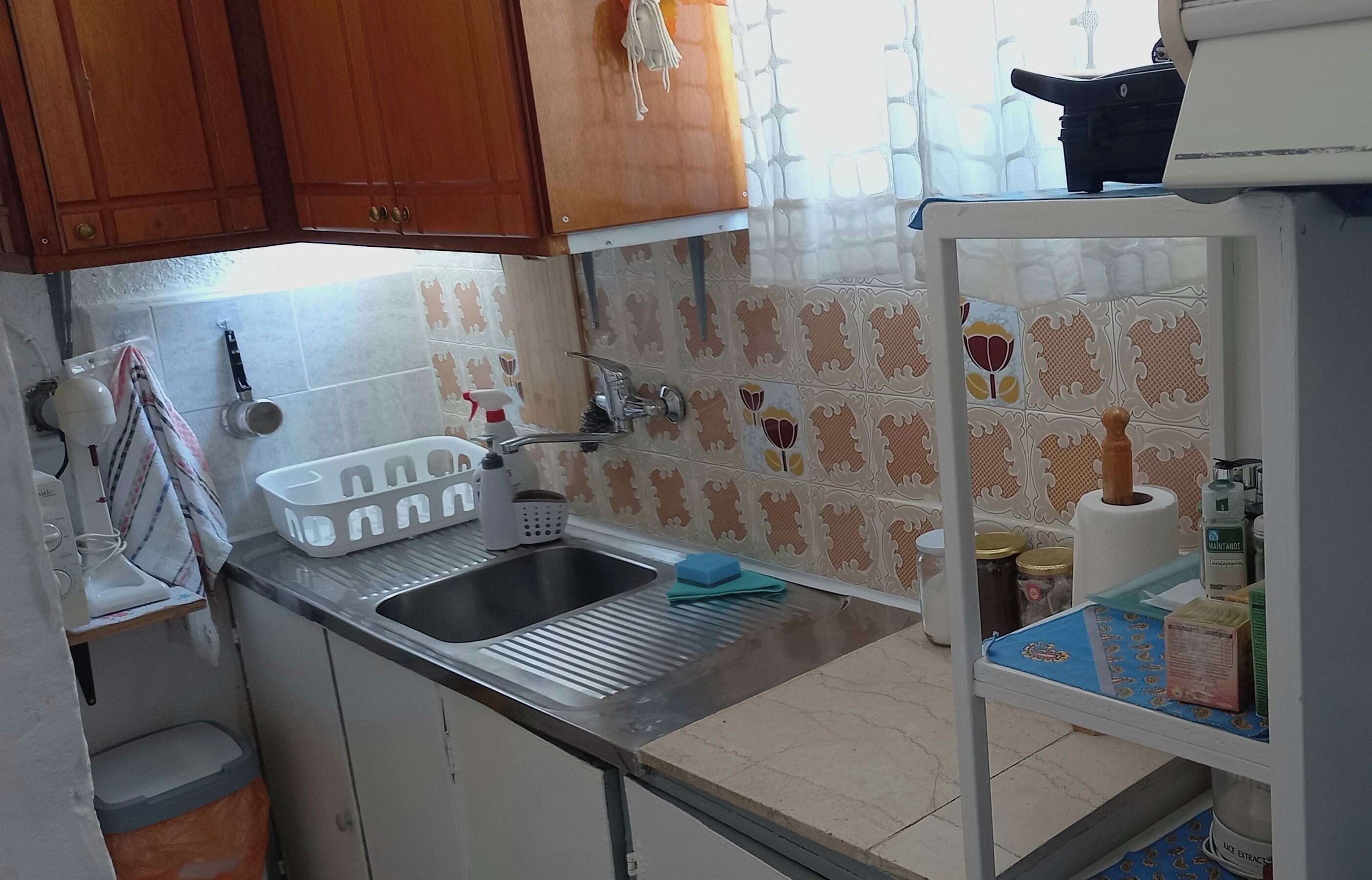A kitchen with wooden cabinets, a stainless steel sink, a dish rack, a blue sponge, and a refrigerator with items on the door.