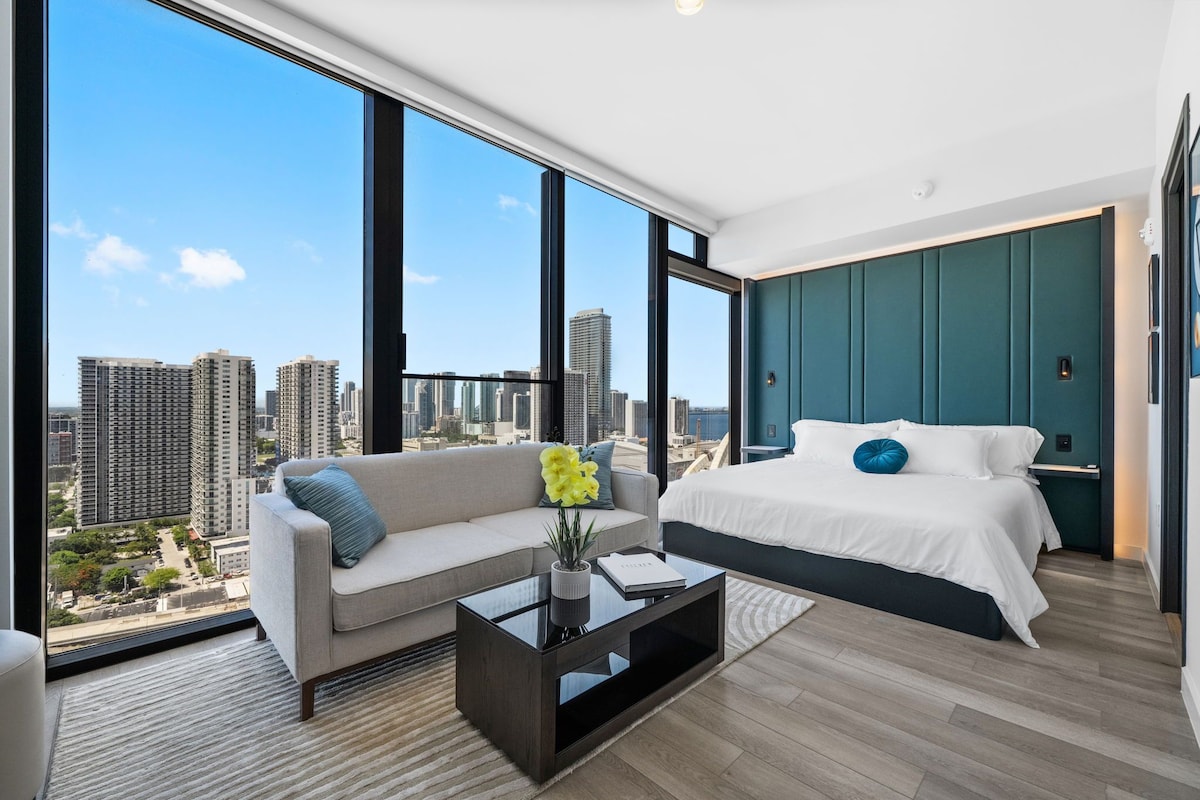 A modern hotel room with a large city view, a bed, a sofa, and a coffee table.
