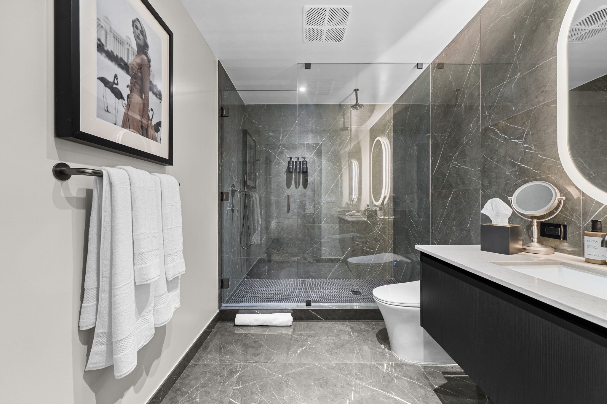 A modern bathroom with a glass-enclosed shower, a toilet, a vanity with a mirror, and a framed picture on the wall.