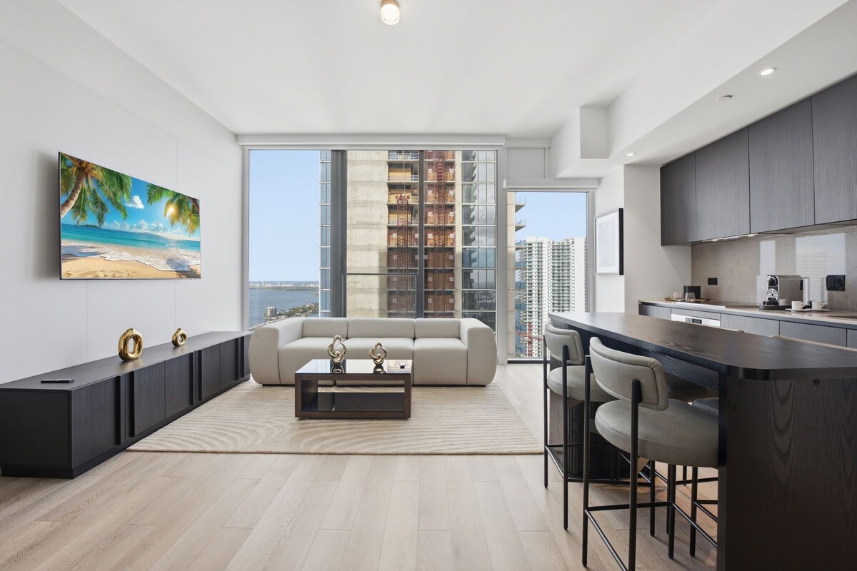 A modern apartment with a living area, kitchen, and a large window overlooking a cityscape.