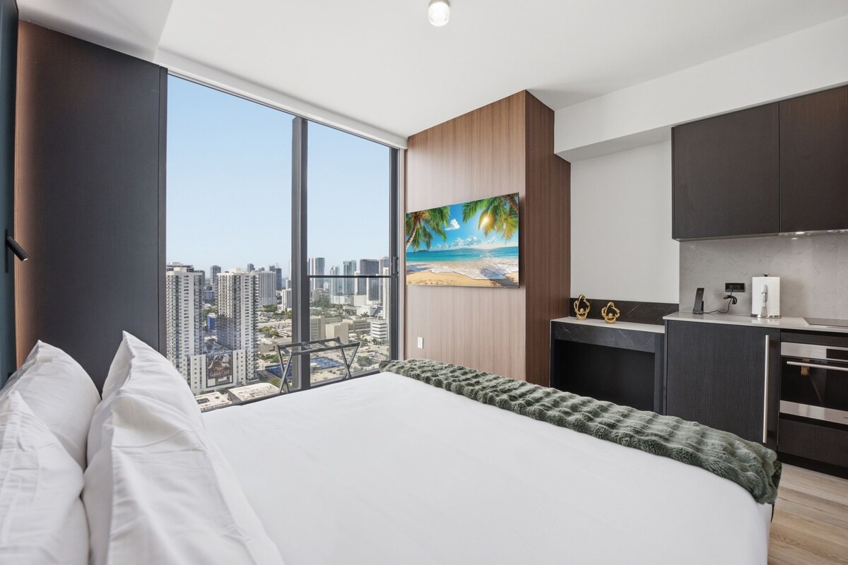 A modern hotel room with a large bed, a city view, a painting of a beach, and a kitchenette.
