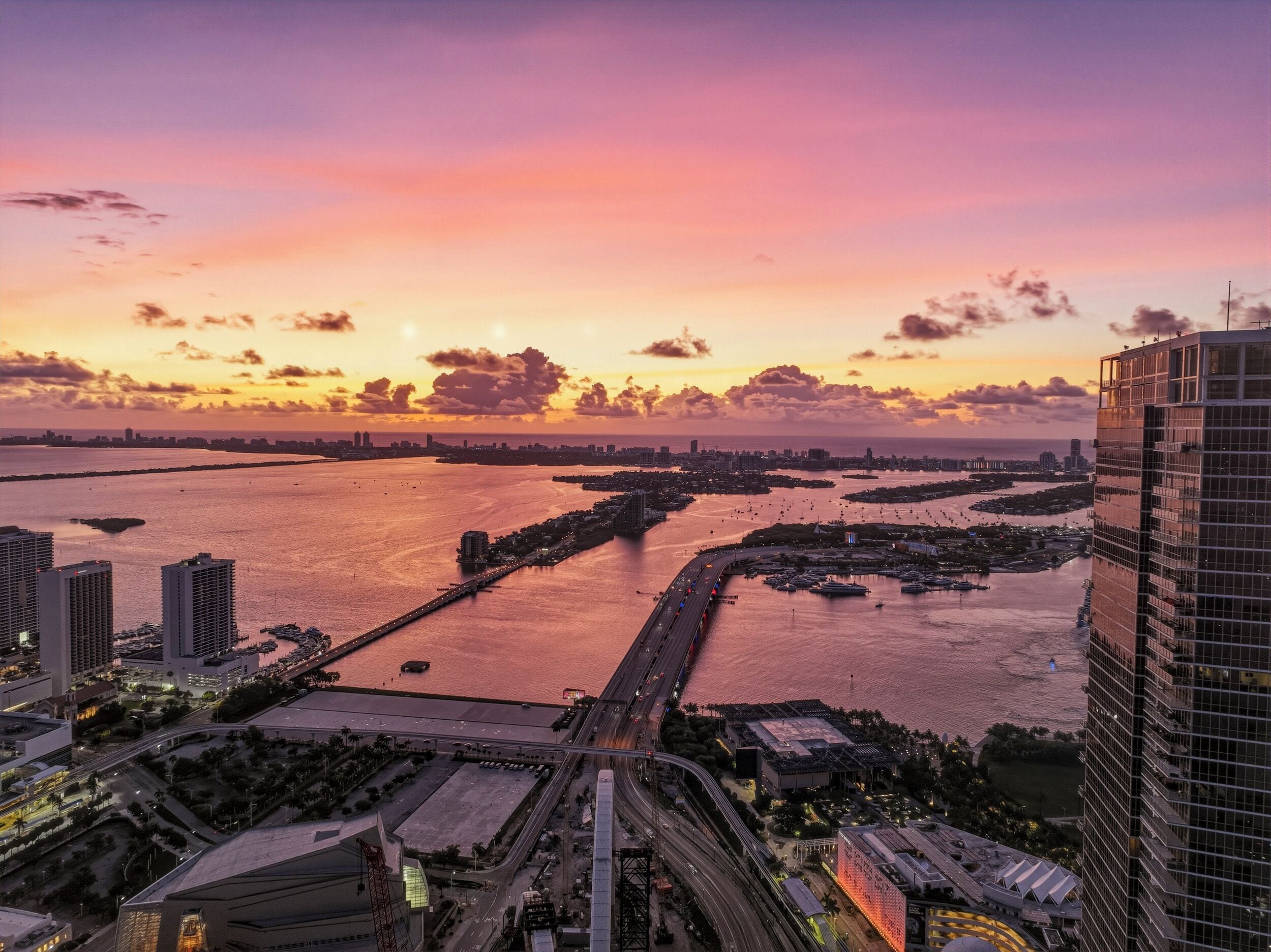 A cityscape with a prominent bridge, high-rise buildings, and a waterfront during sunset.