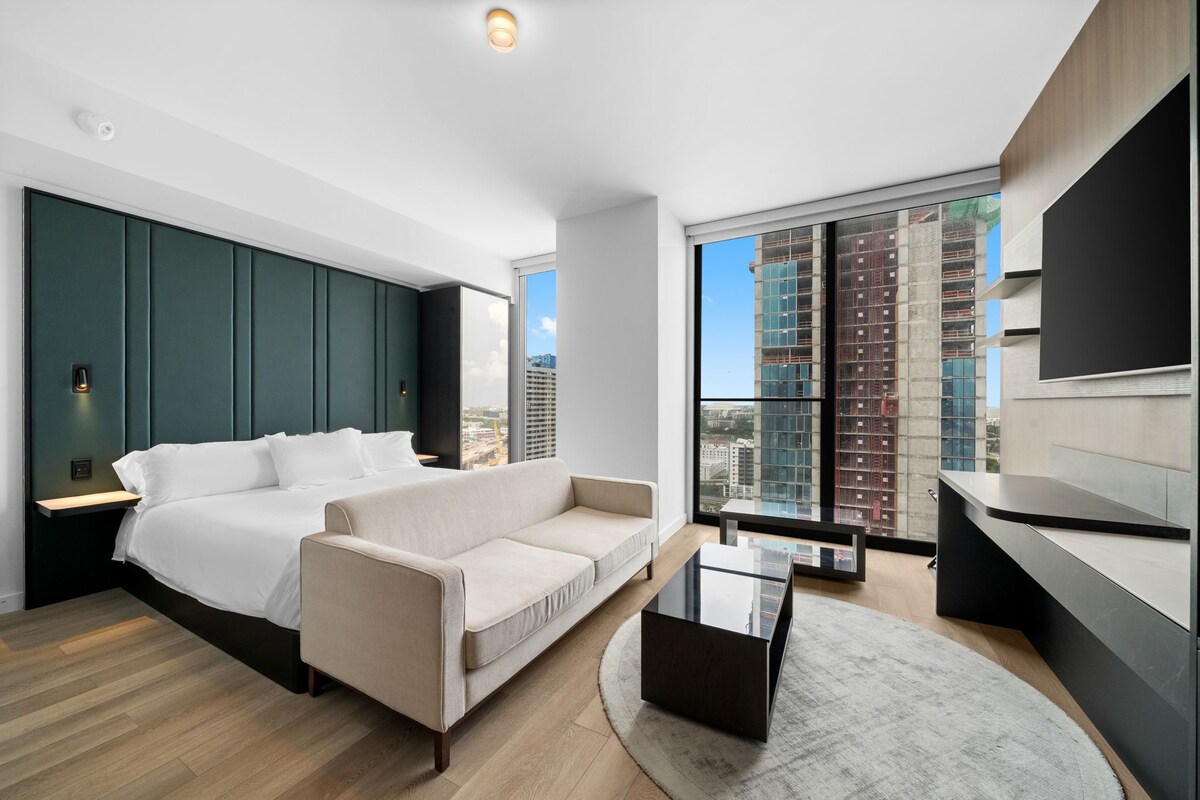 A modern hotel room with a bed, sofa, and a dining area with a view of the city.