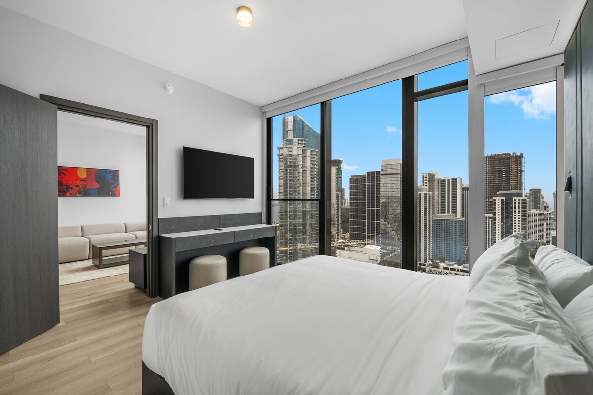 A modern hotel room with a large bed, a flat-screen TV, a seating area with a sofa and chairs, and a city view through large windows.