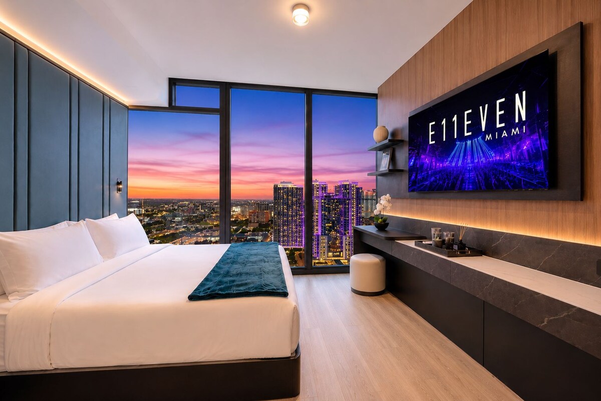 A modern hotel room with a large bed, a TV mounted on the wall, and a city view.