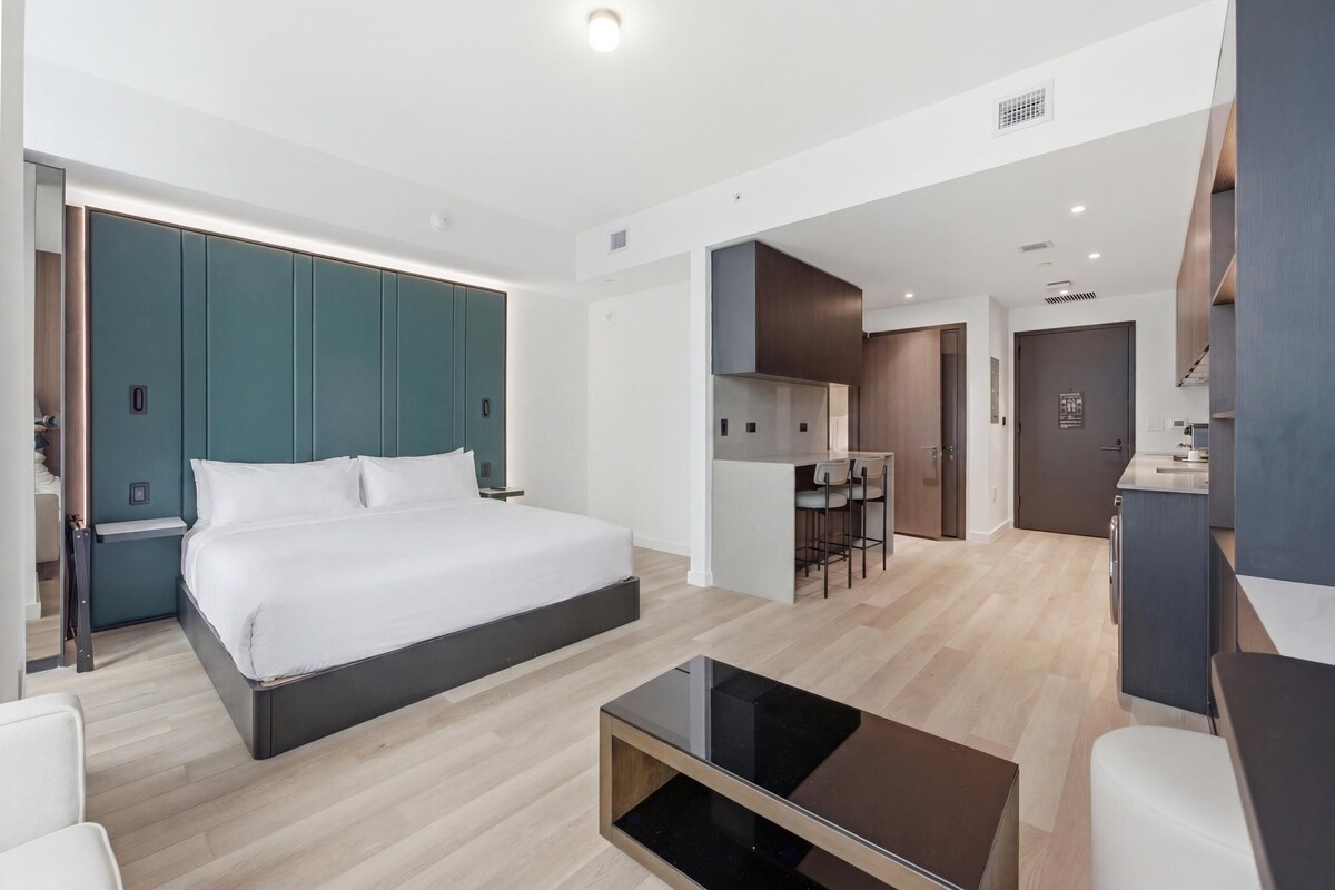 A modern hotel room with a large bed, a desk, and a kitchenette.