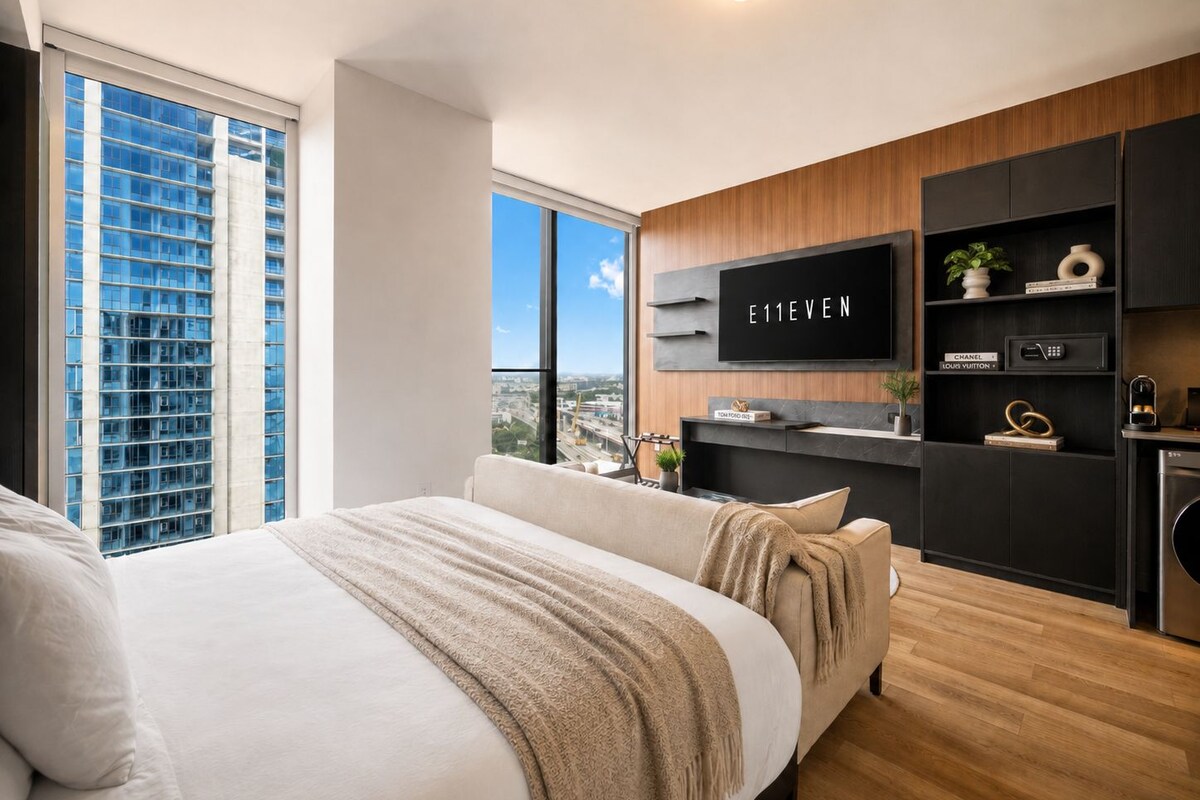 A modern bedroom with a large bed, a TV mounted on the wall, and a city view through the window.