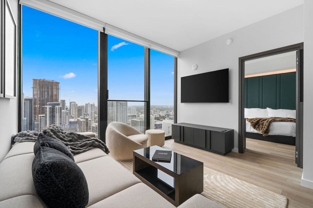 A modern living room with a large window, a sofa, a coffee table, a TV, and a bed with a view of the city skyline.