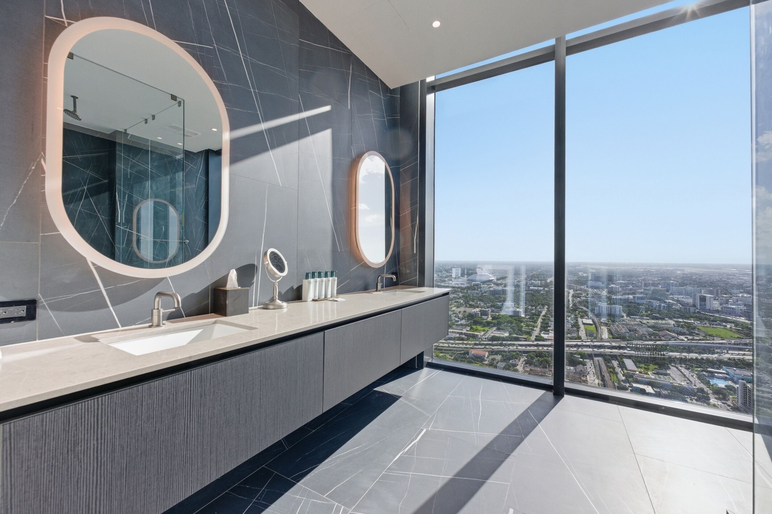 A modern bathroom with a large mirror, two sinks, and a view of a cityscape through large windows.
