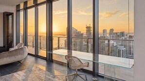 Un interior moderno con ventanas amplias, una mesa de vidrio, una silla acolchada y vistas a un paisaje urbano al atardecer.
