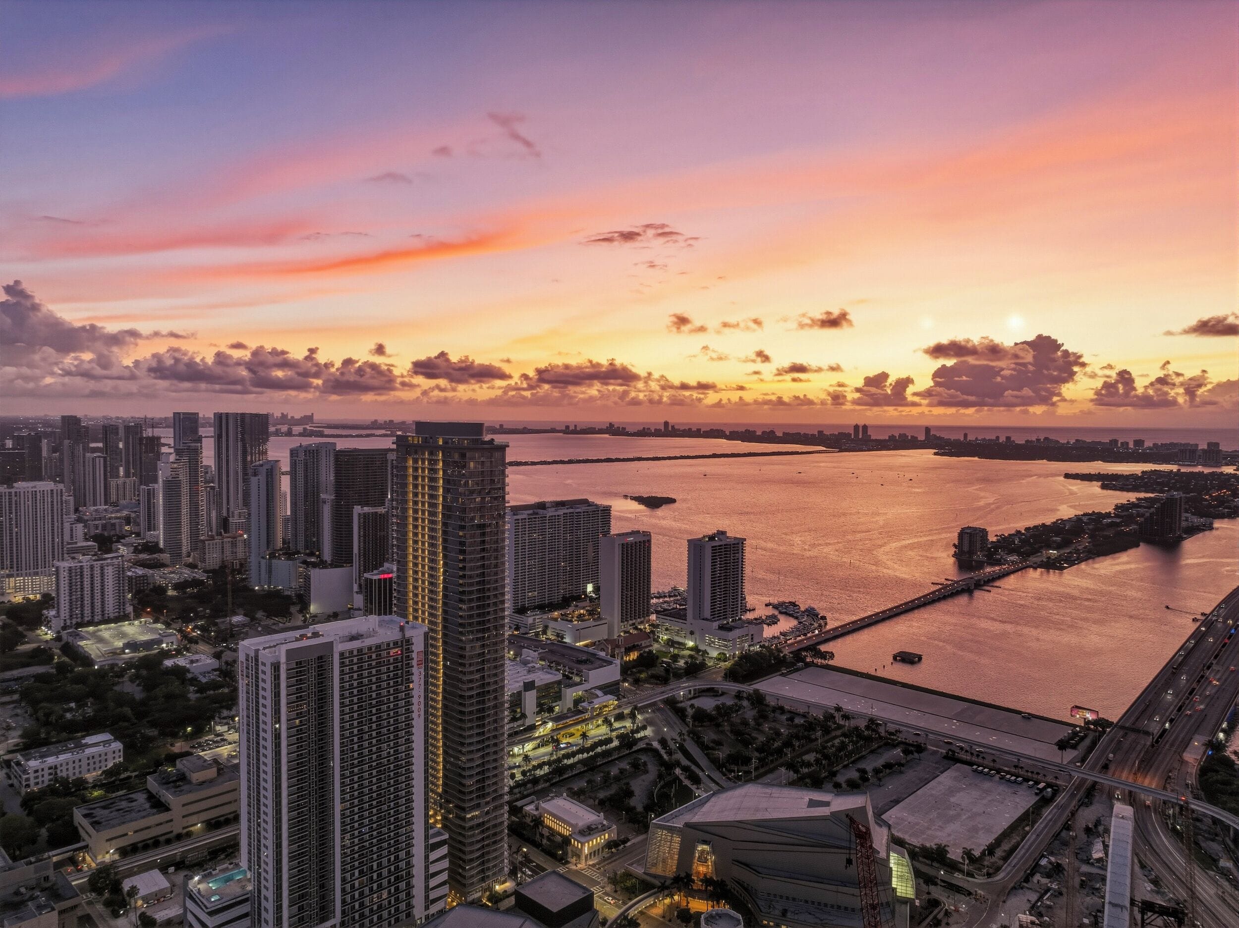 Paisagem urbana com edifícios altos, uma ponte e uma orla durante o pôr do sol.