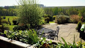 Eine Terrasse mit rundem Tisch und Stühlen, umgeben von Pflanzen und Blick auf eine bewaldete Landschaft.