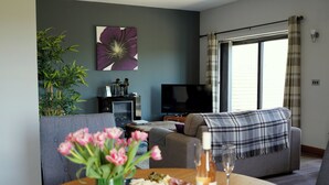 Ein Wohnzimmer mit einer Couch, einem Fernseher, einem Tisch mit Blumen und einem Weinglas sowie einem Fenster mit Vorhängen.