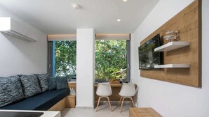 Ein modernes Wohnzimmer mit einer Couch, zwei Sesseln, einem Holztisch, einem Fernseher und einem großen Fenster mit Blick auf Grünflächen.