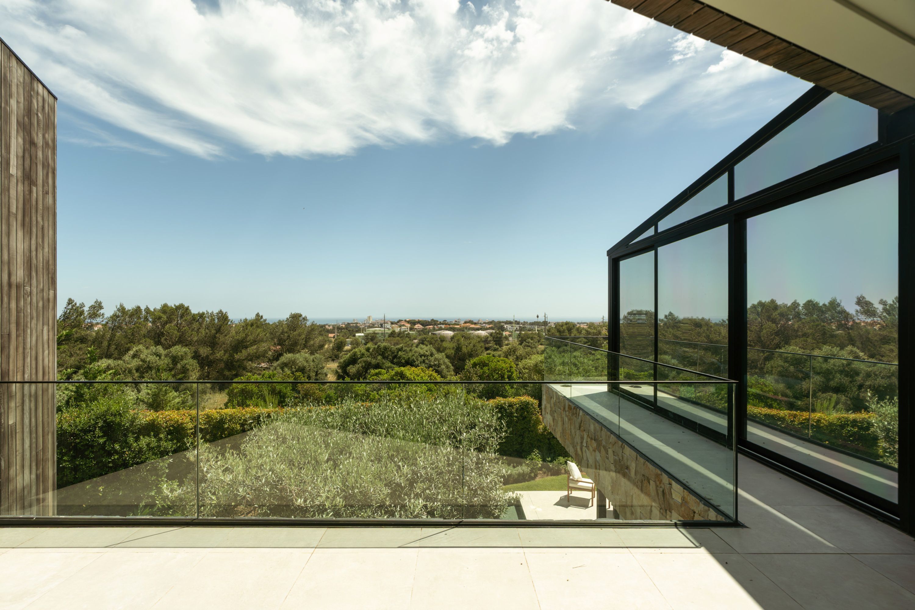 Ein modernes Gebäude mit großen Glasfenstern, einem Balkon mit Blick auf Grünflächen und eine entfernte Landschaft, und einem klaren Himmel.