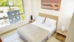 A hotel room with a bed, a nightstand, a bedside table, a window with a view of buildings, and a wall-mounted picture. - Beautiful apartment in the San Gil Plaza shopping center (San Gil)