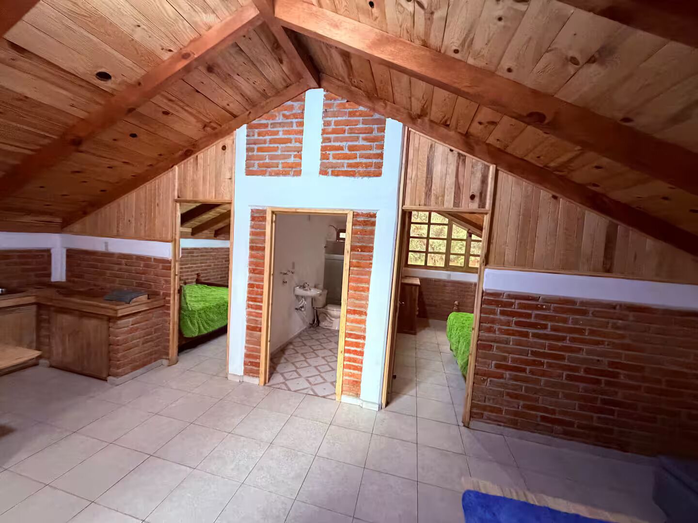 A loft-style room with a sloped ceiling, wooden beams, a brick wall, a small bathroom, and a bed with a green blanket.