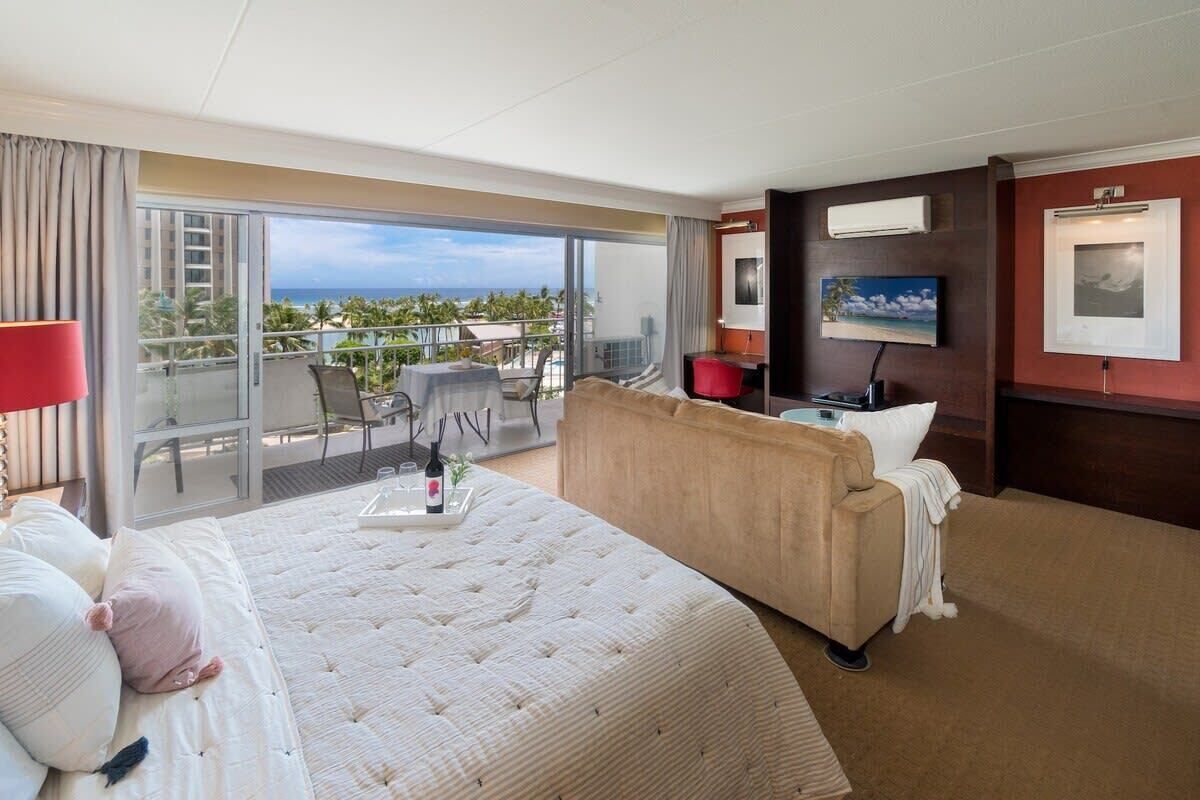Habitación de hotel con una cama grande, un sofá, un balcón con vista al océano y un televisor en la pared.