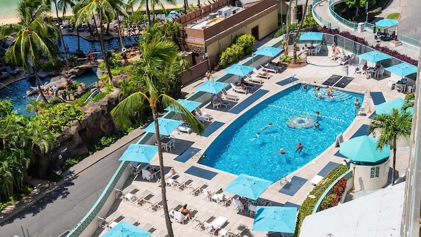 Una gran piscina al aire libre rodeada de sillas de descanso y sombrillas, con palmeras y un edificio al fondo.