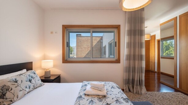 Ein Hotelzimmer mit Bett, Nachttisch, Fenster mit Blick auf ein Gebäude und einer Tür zu einem anderen Raum.