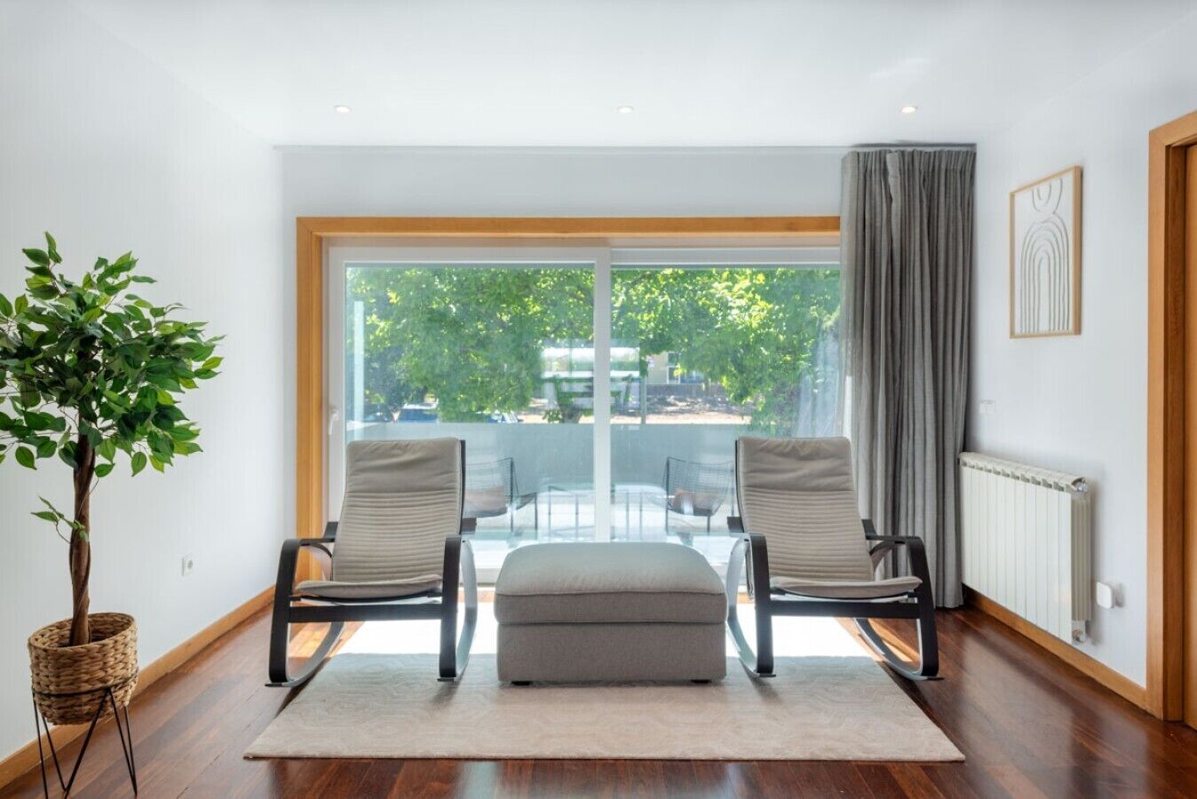A modern living room with a large sliding glass door, two armchairs, a small ottoman, a potted plant, and a radiator.