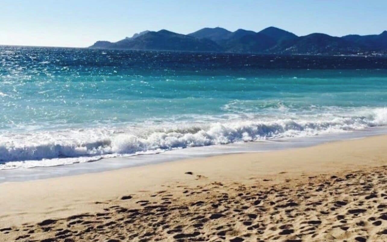 Une plage avec une eau cristalline, des vagues et des montagnes en arrière-plan.