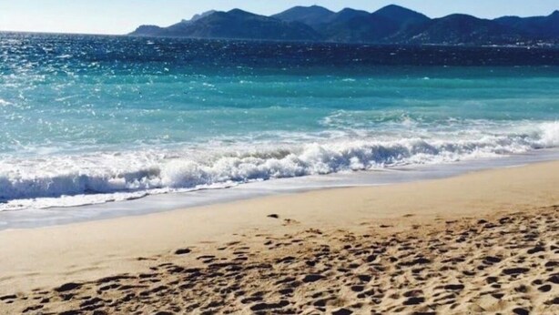 Ein Strand mit klarem Wasser, Wellen und Bergen im Hintergrund.