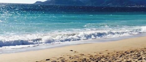 Une plage avec une eau cristalline, des vagues et des montagnes en arrière-plan.