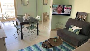 Ein Wohnzimmer mit einer Couch, einem Fernseher, einem Esstisch mit Stühlen und einem kleinen Tisch mit einer Fernbedienung.