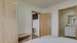 A bedroom with a bed, a wooden wardrobe, a door leading to a bathroom, and a small table with a chair. - Maiata Beach 3 (Madeira)