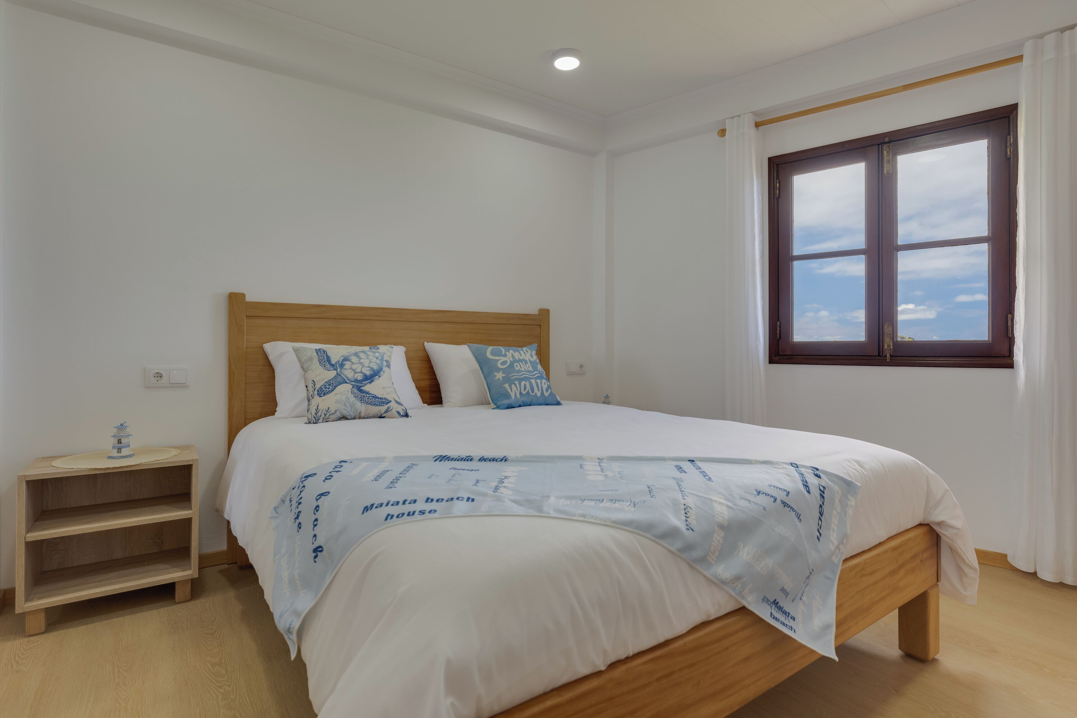 A bedroom with a wooden bed, white bedding, a nightstand, a window with curtains, and a ceiling light.