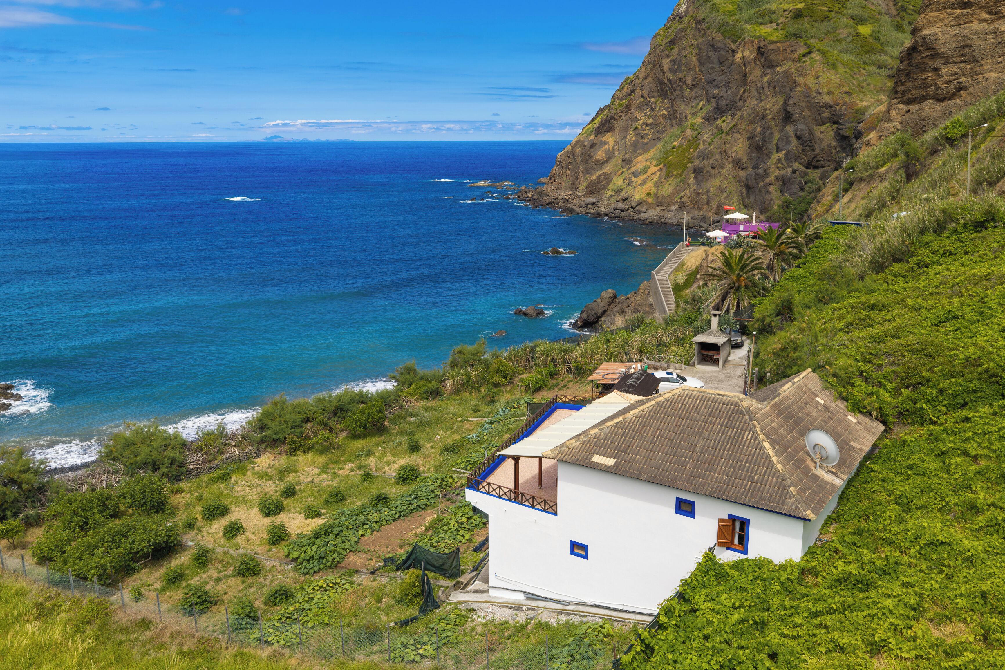 A coastal house with a view of the sea, surrounded by greenery and a rocky cliff.