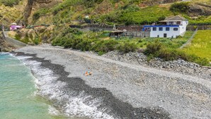 A coastal property with a rocky beach, a white building with blue trim, and a hillside with vegetation. - Maiata Beach 3 (Madeira)