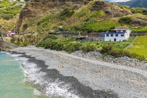 A coastal property with a rocky beach, a white building with blue trim, and a hillside with vegetation. - Maiata Beach 3 (Madeira)