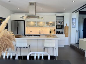 A modern kitchen with white cabinetry, a central island, stainless steel appliances, and a dining area with white chairs. - VillaSerenita Heated Pool, Jacuzzi, Ping Pong (ALATA)