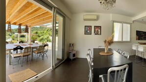 Een moderne keuken met een ingebouwde oven, een eethoek met een grote tafel en stoelen, en uitzicht op een weelderige tuin door een glazen schuifdeur.