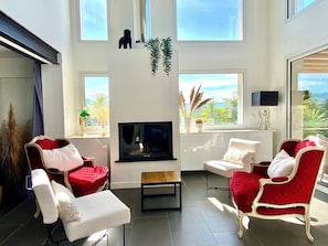 A modern living room with a fireplace, red and white furniture, and large windows offering a view of the outdoors. - VillaSerenita Heated Pool, Jacuzzi, Ping Pong (ALATA)